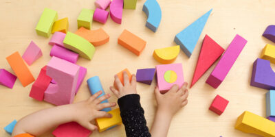 A overhead view of two children manipulating colorful blocks over the flat wooden surface of a table. Their heads are cut by the bottom frame. The child on the left bottom side wears an orange tee-shirt and sticks their two arms to touch a wooden blue and yellow block that resembles a hammer and a cubical pink, green, and yellow block. The child on the right bottom side wears a long sleeve black shirt with white dots and sticks their left arm out to touch an orange wooden block of irregular shape. On the table there are about thirty more blocks of all kinds of sizes, colors, and shapes.