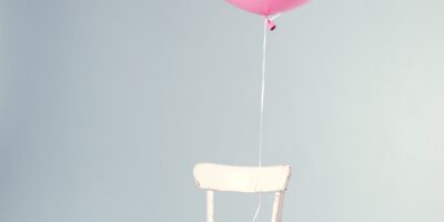 A round pink balloon tethered and floating above tethered to an empty white chair against a grey background