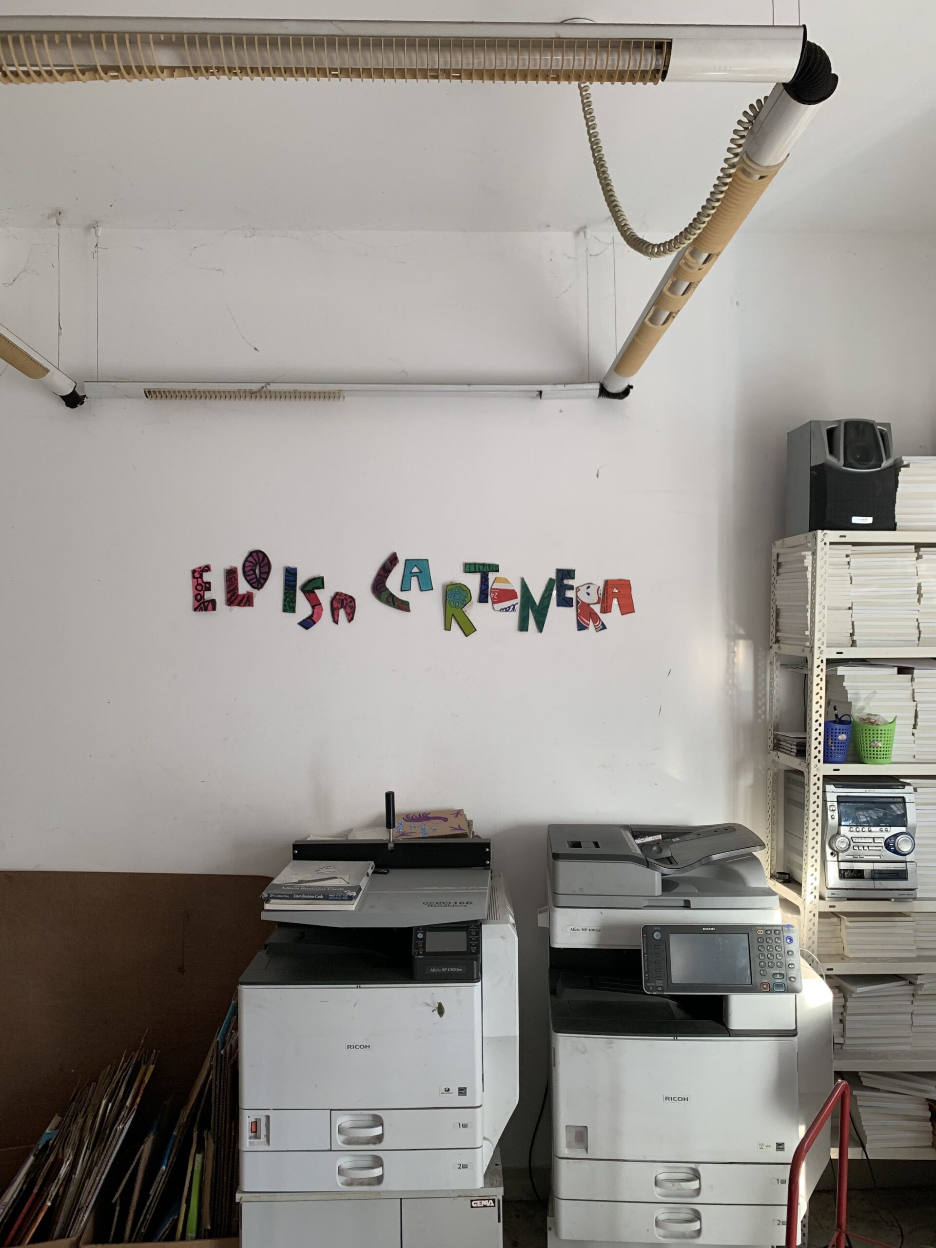 two printers white a white wall above them. Mounted on the wall are colorful cardboard letters that read "Eloisa Cartonera"