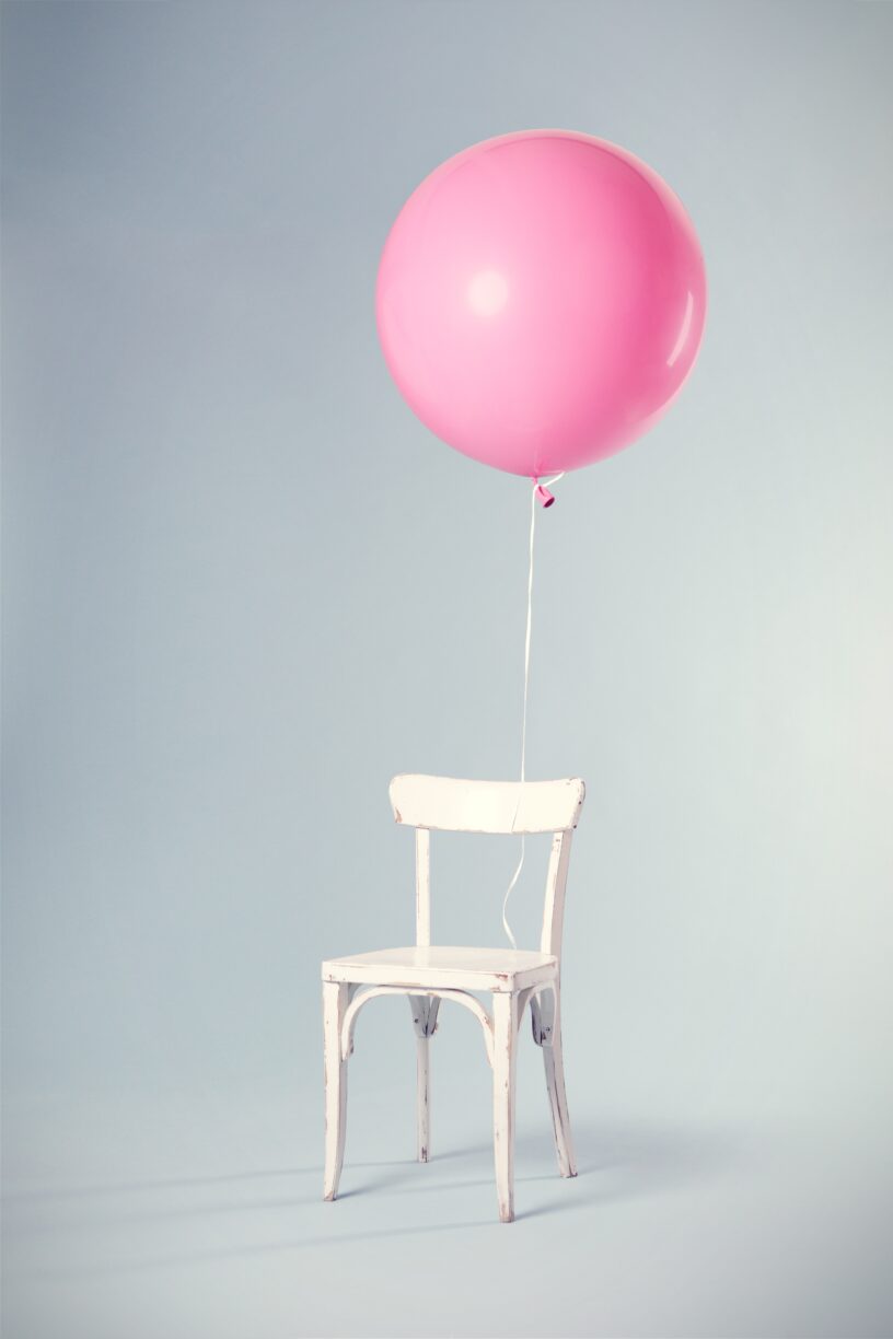 A round pink balloon tethered and floating above tethered to an empty white chair against a grey background