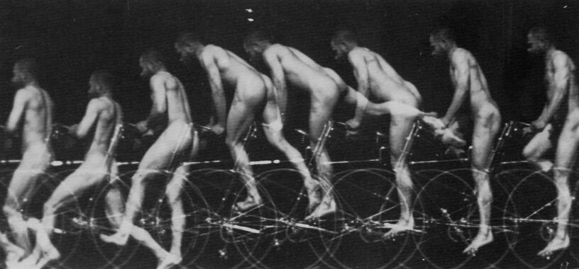 A black and white multilayered and sequential image of a naked bearded person with short hair dismounting a bicycle. The picture is taken from the back. From right to left there are eight superimposed positions that show the motion effect of the person as he lifts his right leg over the bicycle and places his two feet on the ground without letting go of the handlebar.